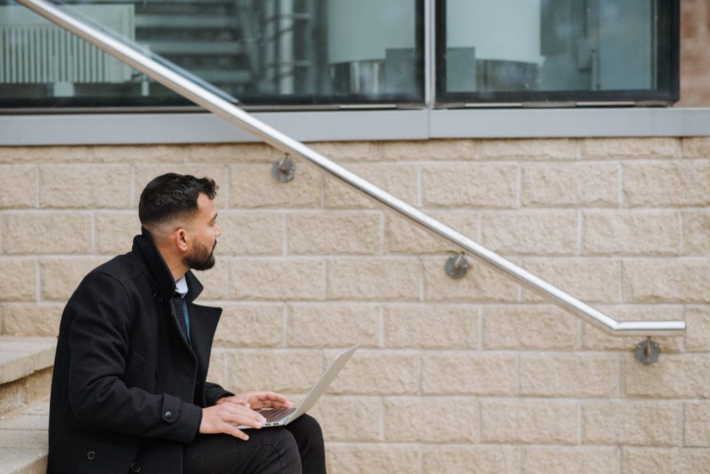 Side view of thoughtful ethnic male manager in formal clothes sitting on street staircase with netbook while looking away