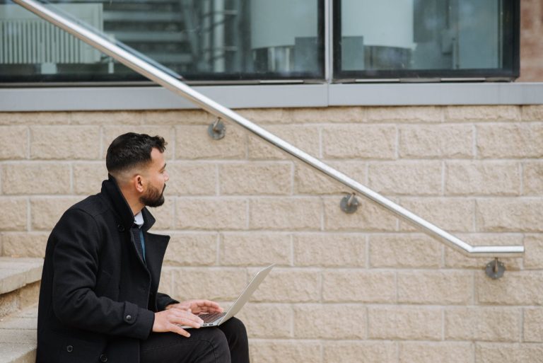 Side view of thoughtful ethnic male manager in formal clothes sitting on street staircase with netbook while looking away