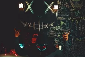 A masked individual stands before a chalkboard filled with neon drawings and cryptic messages, creating a mysterious ambiance.