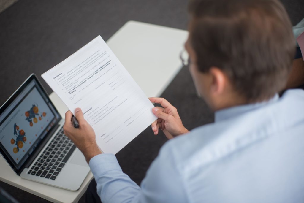 Professional in blue shirt reviewing documents next to laptop displaying charts.