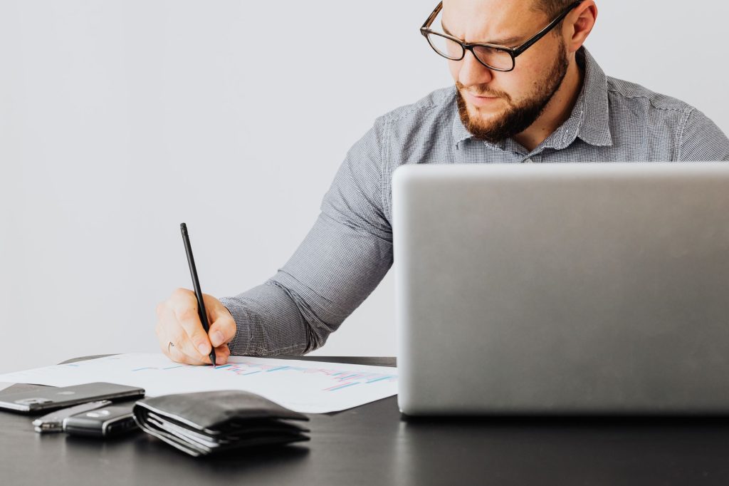 A focused man analyzing financial papers with a laptop in an office setting.