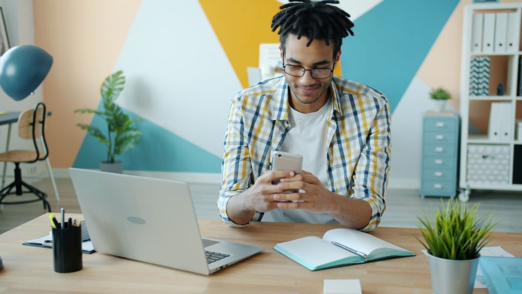 Young adult using a smartphone at a desk with a laptop in a modern office.